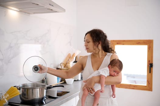 Beautiful curly haired Latin American young adult mother carrying her newborn baby, standing by an electric stove and cooking lunch in a modern home kitchen. People. Family. Lifestyle. Domestic life