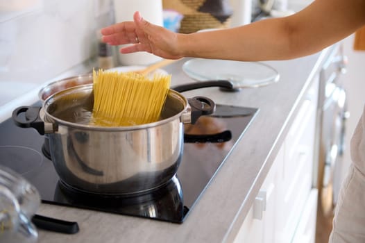 Close-up woman putting Italian pasta spaghetti in a saucepan with a boiling water, standing by an electric stove in modern kitchen.