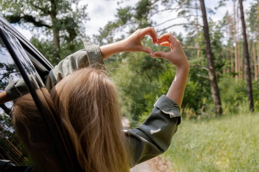Woman riding car holding hand out of window showing heart symbol with her hands. Love and adventure Local traveling concept. Freedom and connection with nature. Sustainable lifestyle roadtrip on weekends to relax and rest