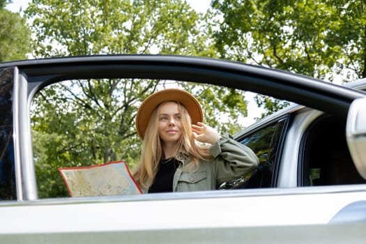 Blonde woman in hat staying next to car door checks the route on map to get to the destination. Young tourist explore local travel making candid real moments. True emotions expressions of getting away and refresh relax on open clean air