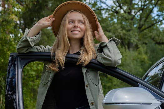 Smiling young woman in hat stoped on road to make a break in driving car. Local solo travel on weekends concept. Exited woman explore freedom outdoors in forest. Unity with nature lifestyle, rest recharge relaxation