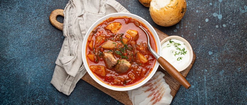 Ukrainian Borscht, red beetroot soup with meat, in white bowl with sour cream, garlic buns Pampushka and salo slices on rustic stone background. Traditional authentic dish of Ukraine