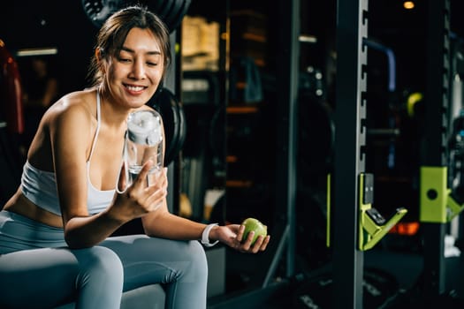 Asian girl holds green apple and water bottle in a gym, reminding viewers of the importance of balanced diet and hydration for achieving healthy lifestyle. Healthy fitness and eating lifestyle concept