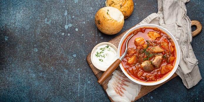 Ukrainian Borscht, red beetroot soup with meat, in white bowl with sour cream, garlic buns Pampushka and salo slices on rustic stone background. Traditional authentic dish of Ukraine, space for text