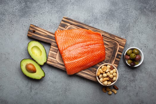 Food sources of healthy unsaturated fat: fresh raw salmon fillet, avocado, olives, nuts on cutting board on rustic stone background top view. Omega 3 and oil products, healthy nutrition and keto diet