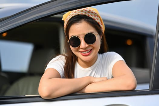 Portrait of female traveler leaning out from car window and smiling at camera. Travel and vacation concept.