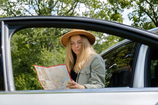 Blonde woman in hat staying next to car door checks the route on map to get to the destination. Young tourist explore local travel making candid real moments. True emotions expressions of getting away and refresh relax on open clean air