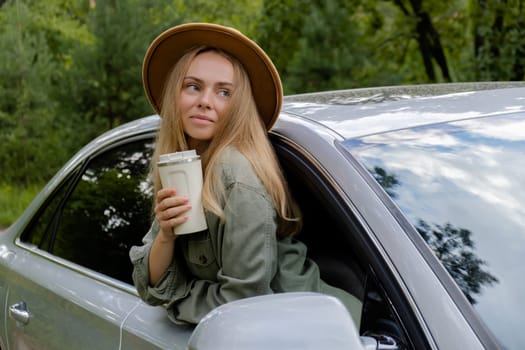 Blonde woman sticking head out of windshield car and drink coffee or tea from reusable mug. Young tourist explore local travel making candid real moments. Refuse reuse recycle zero waste concept. Responsible traveling reduce carbon footprint sustainable lifestyle