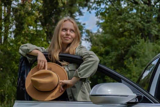 Blonde woman in hat staying next to car door. Young tourist explore local travel making candid real moments. True emotions expressions of getting away and refresh relax on open clean air