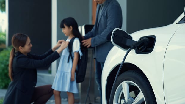 Progressive young parents and daughter with electric vehicle and home charging station. Green and clean energy from electric vehicles for healthy environment. Eco power from renewable source.