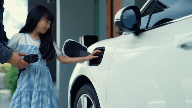 Progressive father and daughter installing plug from home charging station for electric vehicle. Future eco-friendly car with EV cars powered by renewable source of clean energy.