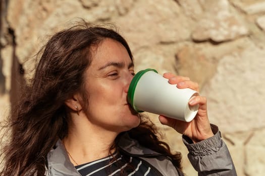 A middle-aged woman drinks coffee from a paper cup outside on a sunny autumn day. High quality 4k footage