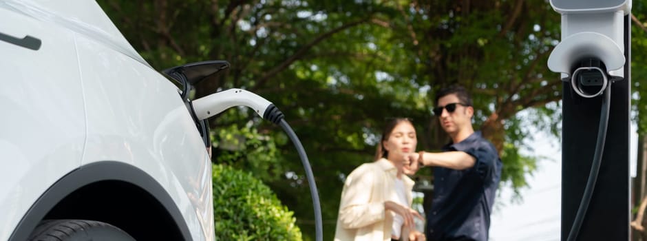 Young couple recharge electric car battery from charging station in green city park in springtime. Rechargeable EV car for sustainable environmental friendly urban travel lifestyle. Panorama Expedient