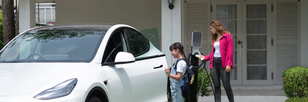 Happy little young girl learn about eco-friendly and energy sustainability as she help her mother recharge electric vehicle from home EV charging station. EV car and modern family. Panorama Synchronos