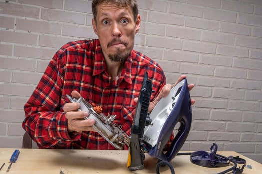 a funny and annoyed grief-stricken master repairs an electric iron. the master repairs the iron in the workshop on the table using tools. Disassembled electric iron.