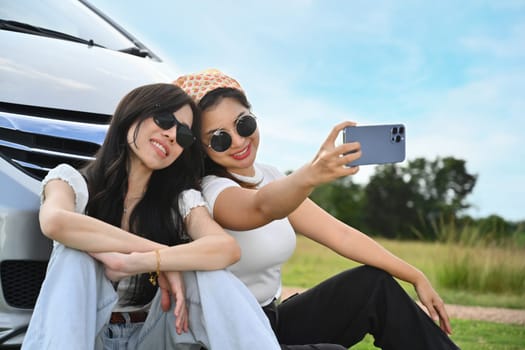 Happy female friends taking selfies with smartphone siting in front of car on meadow.