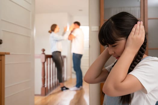 Stressed and unhappy young girl huddle in corner, cover her ears blocking sound of her parent arguing in background. Domestic violence at home and traumatic childhood develop to depression. Synchronos