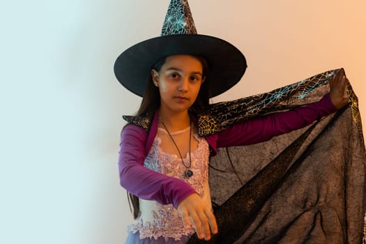 Happy scary Halloween concept: little witch girl playing paranormal Halloween role and doing scary look on a yellow background. Kid scaring and making Boo in carnival witch costume or wizard costume.