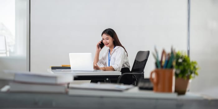 Happy young asian businesswoman talk on the mobile phone and smile while sitting at working place in office.