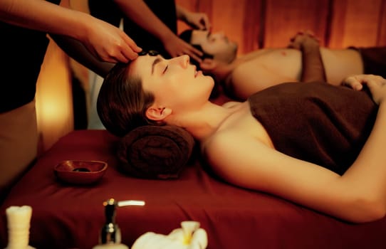 Caucasian couple enjoying relaxing anti-stress head massage with hot stone and pampering facial beauty skin recreation leisure in warm candle light ambient salon spa in resort or hotel. Quiescent