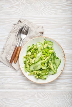 Healthy vegan green avocado salad bowl with sliced cucumbers, edamame beans, olive oil and herbs on ceramic plate top view on white wooden rustic table background