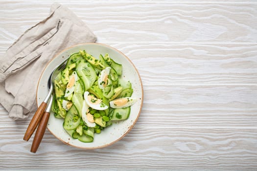 Healthy green avocado salad bowl with boiled eggs, sliced cucumbers, edamame beans, olive oil and herbs on ceramic plate top view on white wooden rustic table background. Space for text