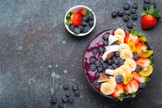 Healthy summer acai smoothie bowl with chia seeds, fresh banana, strawberry, blueberry, cocos, kiwi top view on rustic concrete background with spoon. Space for text