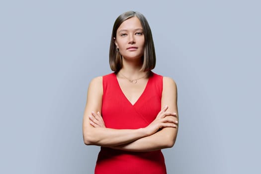 Portrait of young confident woman in red on grey studio background. Successful fashionable female with crossed arms looking at camera. Business, work, services, education, fashion beauty professions