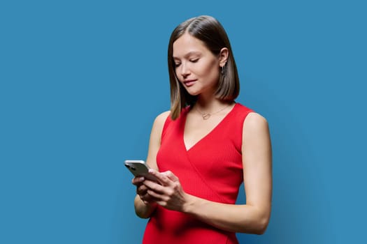 Young woman with smartphone in hands on blue studio background. Serious female looking at phone screen texting reading. Using mobile applications for work business shopping banking study leisure