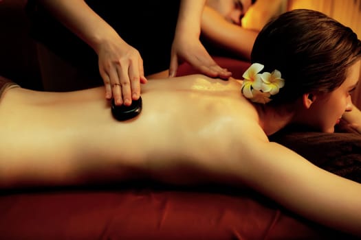 Hot stone massage at spa salon in luxury resort with warm candle light, blissful couple customer enjoying spa basalt stone massage glide over body with soothing warmth. Quiescent