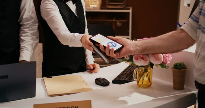 Elderly man makes nfc transaction with mobile phone at pos terminal, paying for accommodation at five star resort. Tourist arriving at front desk doing check in for holiday. Close up. Handheld shot.