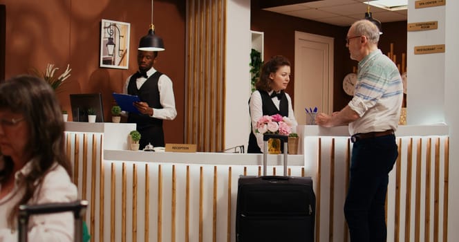 Senior man entering hotel lobby with luggage, asking for concierge services to check in. Receptionist woman ensuring relaxing stay for old tourist at all inclusive holiday resort, tourism industry.
