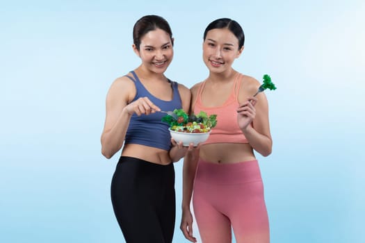 Two young sporty Asian women in sportswear holding salad bowl fill with fruit and vegetable. Natural youthful and fit body lifestyle people with balance nutrition on isolated background. Vigorous