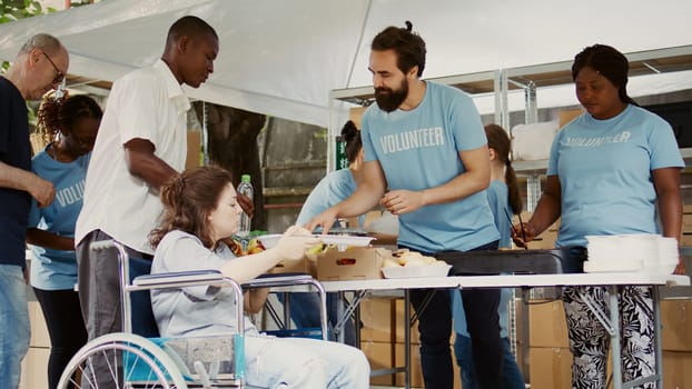 Helpful guy volunteer offers free lunch to the poor disabled woman in need. Multiracial charity workers at an outdoor food bank provide the handicapped and less fortunate with hunger relief.