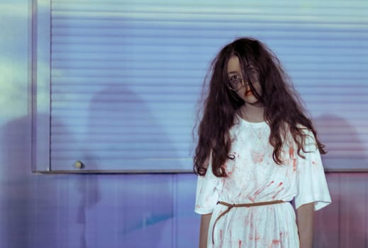 Portrait of a beautiful caucasian teenage girl in glasses, long flowing hair in a bloody white dress with a belt standing on the right against a background of a pale neon wall with copy space on the left, close-up side view.