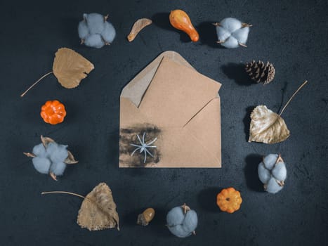 Kraft envelope with a sheet with a place for text lies in the middle on a black background, and around is a decor of leaves, decorative pumpkins, cotton, cobwebs and a spider. Flat lay close up.