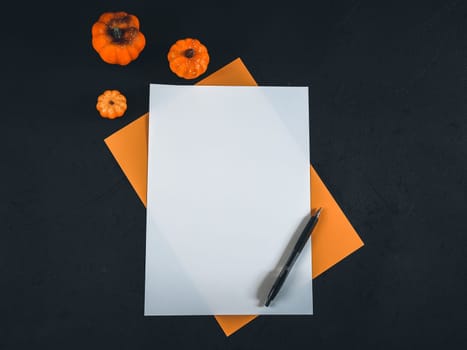 Two blank sheets, white and orange, lie vertically in the middle against a black background with three decorative pumpkins on the left and a pen on the right. Flat lay close up.