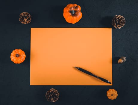 One orange empty leaf, white and orange, lie horizontally in the middle against a black background with decorative pumpkins, pine cones and a pen. Flat lay close up.