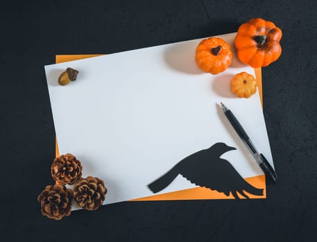 Two empty leaves, white and orange, lie horizontally in the middle against a black background with a paper crow, decorative pumpkins, pine cones and a handle on the sides. Flat lay close up.