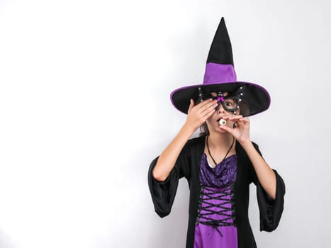 Portrait of a caucasian girl in a witch costume with a halloween hat holds candy near her mouth with one hand, and closes her eye with the other, stands on the right against a white wall with little copy space on the left, close-up side view. Halloween concept, Halloween celebration.