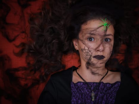 Portrait of a beautiful caucasian brunette girl with brown eyes in a witch's dress with a cobweb on her face and a spider in hair looks directly into the camera while lying on an orange-red sofa, close-up top view. Halloween concept,Halloween celebration.