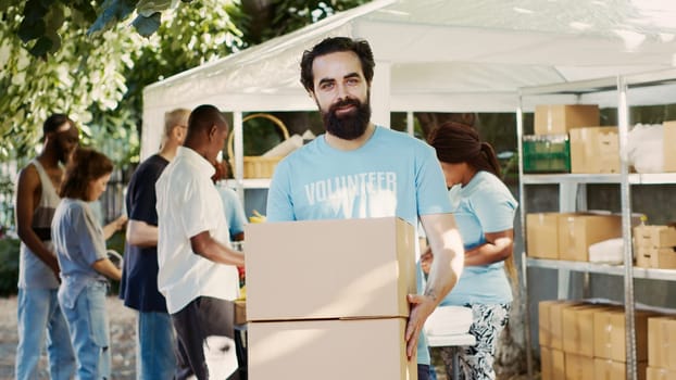 Generous man volunteering at neighborhood food drive, distributing non-perishable rations to the hungry and homeless. While holding donation box, caucasian charity worker looks at camera. Tripod shot.