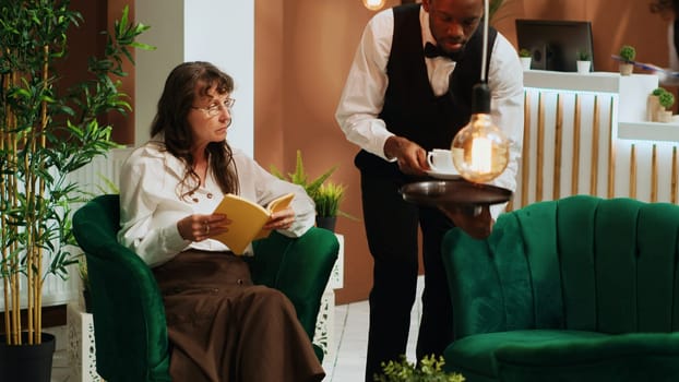 Bellboy serving coffee to woman traveller waiting in lounge area, senior person enjoying drink and reading literature book at hotel. Employee providing all inclusive services. Retirement holiday.