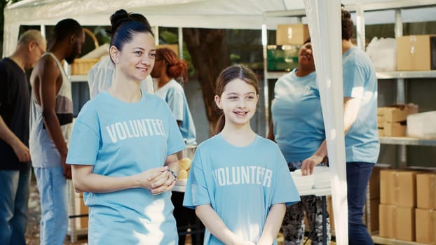 Side-view portrait of a mother and daughter in support of a food bank outside campaign to fight hunger. Two caucasian females in blue volunteer t-shirts are prepared to assist the homeless people.