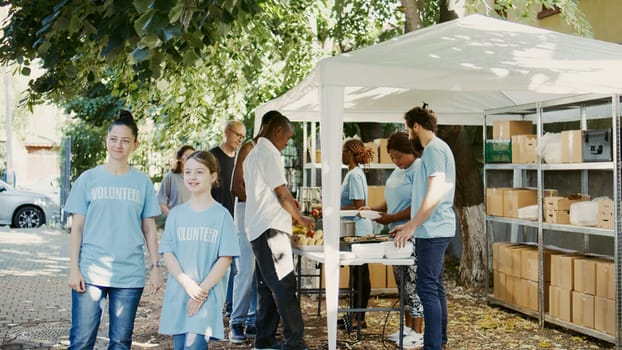 Caucasian mother and her daughter taking part in a hunger relief initiative at a food bank. Multiethnic volunteers in blue t-shirts outdoors ready to help the need and homeless people. Slow-mo.