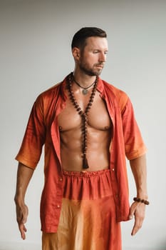 A muscular man with a naked torso in orange clothes stands in a room on a light background.