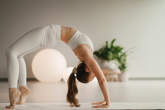 A girl in white clothes does yoga standing on the bridge on a mat indoors.