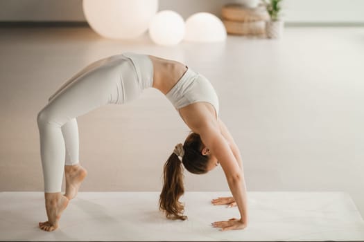 A girl in white clothes does yoga standing on the bridge on a mat indoors.