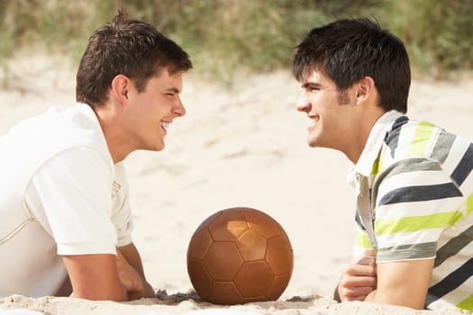 Two Young Men Relaxing On Beach With Football Together