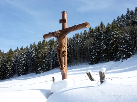 Wood sculpture of Jesus on the cross in front of many fir trees by winter